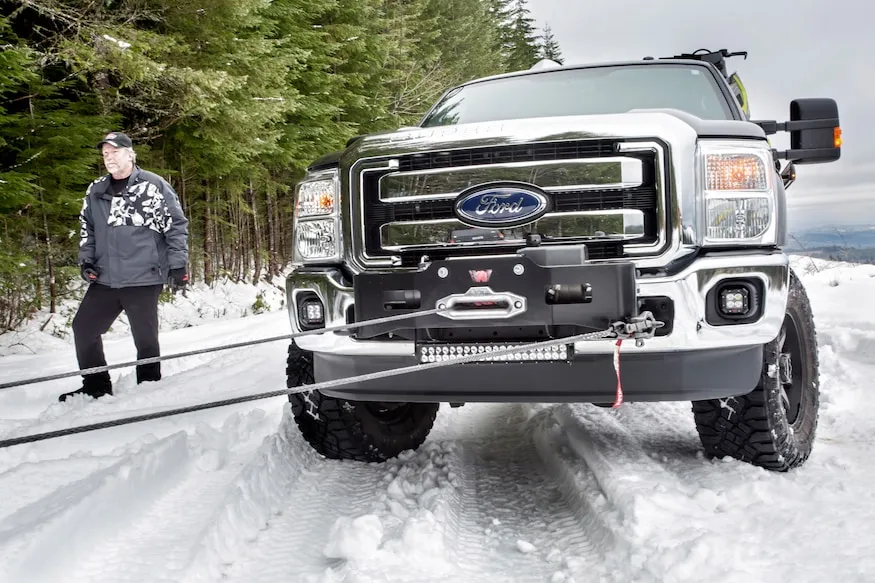 A man driving a truck pulls a snow plow through a snowy road.